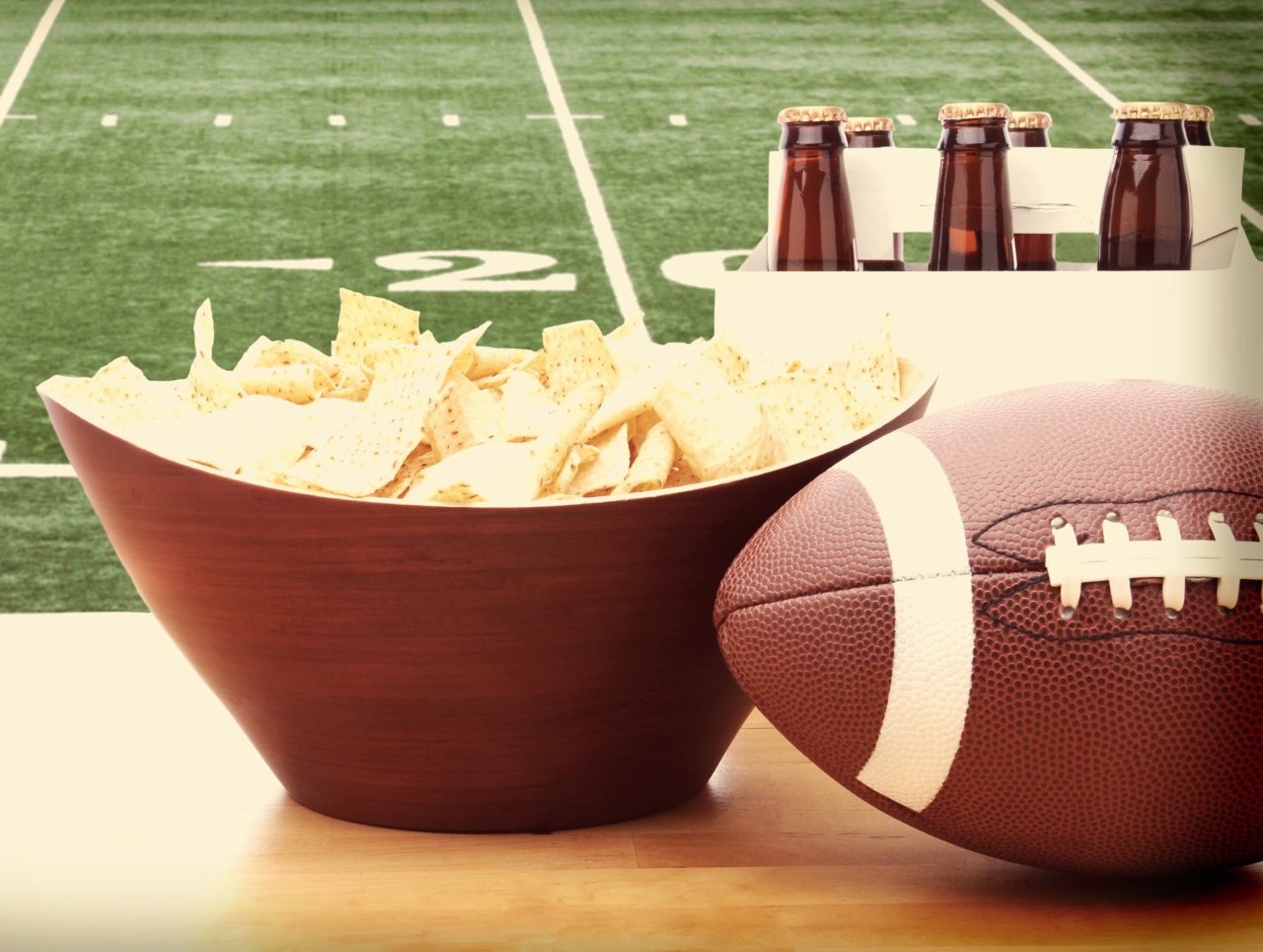 Chips, football and Six Pack of Beer on a table in front of a big screen TV with a Football field. Great for Super Bowl themed projects. Horizontal format with instagram effect applied.