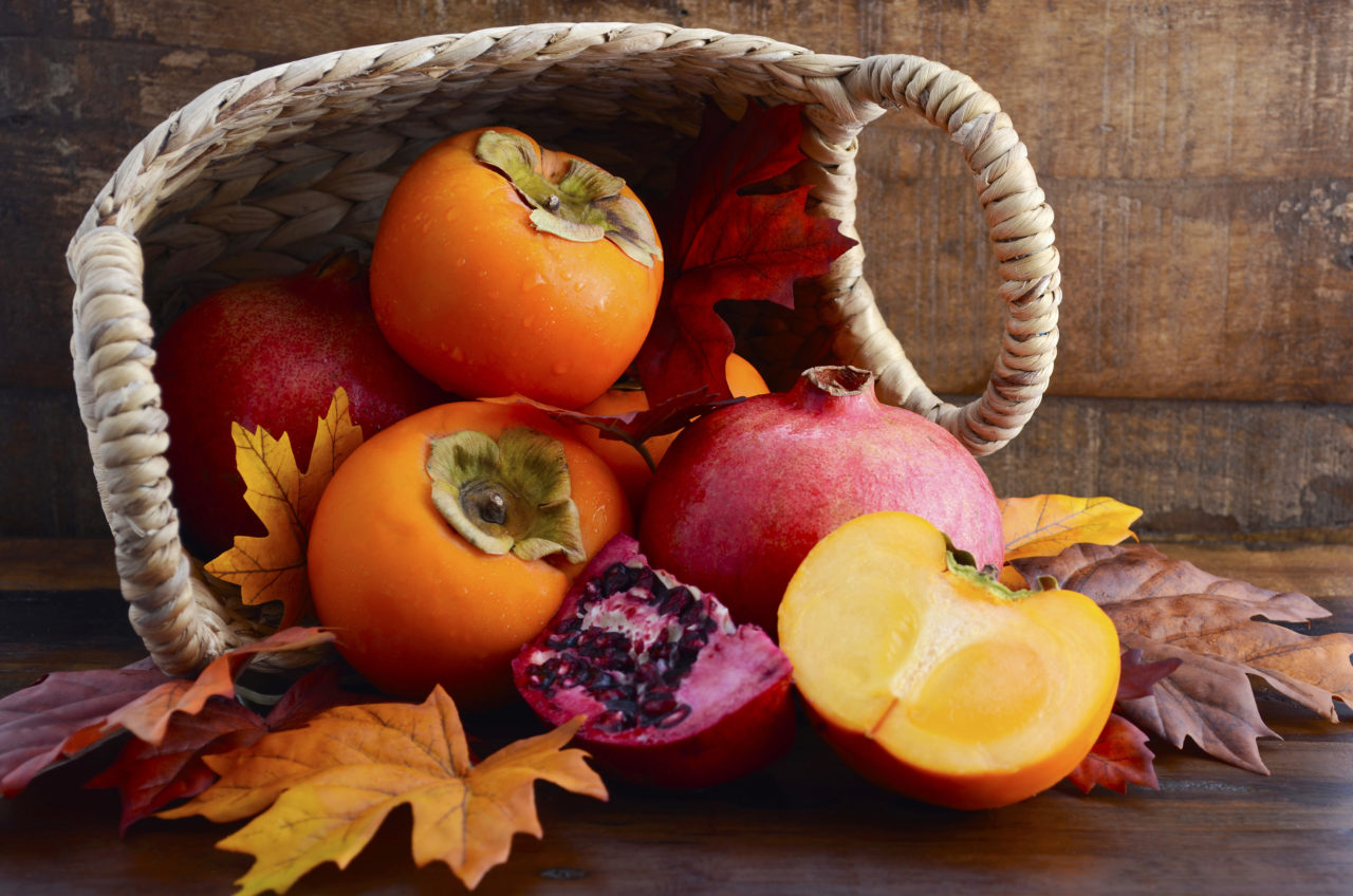Basket of fresh persimmons and pomegranates with cut slices on Autumn Fall leaves against a dark wood vintage background.
